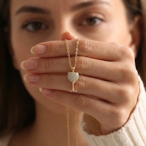 May include: A gold-colored necklace featuring a pendant shaped like a wine glass. The glass portion of the pendant is encrusted with small, sparkling stones. The necklace is held up against a blurred background.