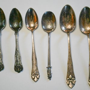 May include: Six antique silver spoons with varying designs. The spoons have oval bowls and ornate handles, some with floral or scrollwork patterns. The spoons are arranged in a row, showcasing their different styles and levels of detail.