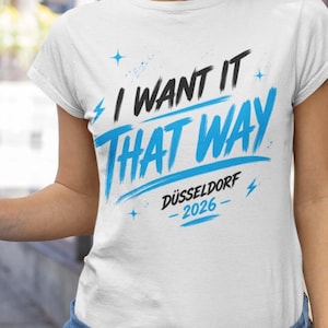 Puede incluir: Una camiseta blanca de manga corta con el texto en negro y azul "I WANT IT THAT WAY" y "DÜSSELDORF 2026" impreso debajo, decorada con pequeños gráficos de estrellas y rayos azules en la parte frontal de la prenda.