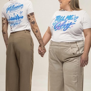 Könnte beinhalten: Zwei weiße T-Shirts mit kurzen Ärmeln. Ein Shirt zeigt den blauen Schriftzug "INTO THE Millennium HOMECOMING DÜSSELDORF 2026" mit einer Stadtsilhouette. Das andere Shirt zeigt den blauen Schriftzug "TELL ME Why" mit kleinen blauen Herz- und Sternmotiven.