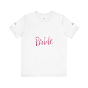 Può includere: T-shirt bianca con la scritta "Bride" in rosa acquerello, ornata da piccoli dettagli di corona e cuore. La parola "Bride" è stampata anche sulle maniche. Un design semplice ed elegante per abbigliamento da sposa.