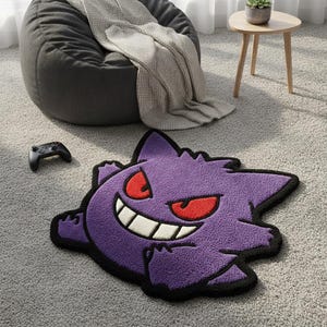 Op de afbeelding: Een paars, getuft vloerkleed in de vorm van het personage Gengar, met een brede witte grijns, scherpe rode ogen en een dikke zwarte rand. Het kleed heeft een pluche textuur en is ontworpen naar het uiterlijk van het bekende spook-type wezen.