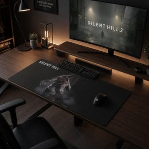 Op de afbeelding: Een donkere bureau-opstelling met een computermonitor met "SILENT HILL 2". Een zwarte muismat met "SILENT HILL" tekst en een afbeelding ligt op het bureau. Een toetsenbord, muis en bureaulamp zijn ook zichtbaar.