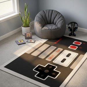 Puede incluir: Una alfombra rectangular con un diseño gráfico en negro, blanco y rojo inspirado en un mando de videojuego clásico, que incluye una cruceta, botones Select y Start, y botones A y B con el logotipo de Nintendo impreso en la superficie.
