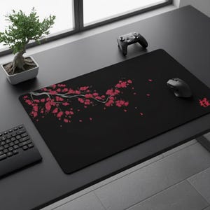 Op de afbeelding: Zwart rechthoekig muismat met een rood bloemenontwerp en Japanse karakters. Een zwarte muis en een gamecontroller liggen op het bureau. Een kleine bonsai en een toetsenbord zijn ook zichtbaar.
