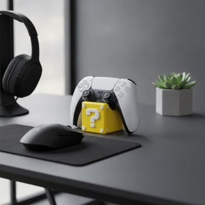 Op de afbeelding: Een wit en zwart videospelcontroller rustend op een gele kubusvormige standaard met een wit vraagteken-symbool. De standaard staat op een donkergrijs oppervlak naast een zwarte computermuis, een headset en een kleine vetplant.