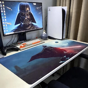 Könnte beinhalten: Ein Computertisch-Setup mit einem Monitor, der Darth Vader zeigt, einer weißen PlayStation 5-Konsole und einem langen Mauspad mit Star Wars-Motiv. Das Mauspad zeigt Darth Vader mit einem roten Lichtschwert vor einem dunkelblauen Hintergrund.