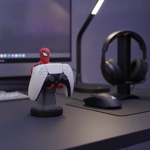May include: A red and blue Spider-Man figurine acting as a controller stand, holding a white and black wireless game controller. The figure stands on a gray pedestal base, designed to securely display the gaming peripheral on a desk surface.