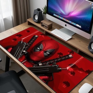 May include: A rectangular red mousepad featuring the character Deadpool with two guns and a katana. The mousepad has a black border and is on a wooden desk with a computer and speakers.
