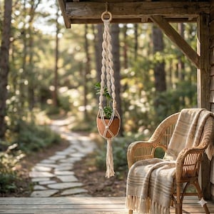 May include: A cream-coloured macrame plant holder with a small potted plant suspended from a wooden beam. A wicker chair with a blanket is in the foreground, with a stone path and trees in the background. The holder has a tassel.