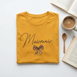 Op de afbeelding: Mosterdgeel T-shirt met het woord "Mawmaw" in een sierlijk lettertype en een luipaardprint strik. Het shirt is op een witte ondergrond gevouwen, met een kop koffie en boeken op de achtergrond.