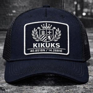 Può includere: Cappellino da camionista blu navy con retro in rete e visiera curva. La parte anteriore presenta una toppa rettangolare bianca con la scritta "KIKUKS", coordinate e un stemma. Un accessorio classico.