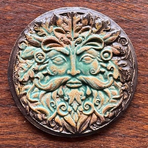 May include: A round, ceramic Green Man face with a teal and brown color scheme. The face has a detailed beard, mustache, and leafy crown. The Green Man is a symbol of rebirth and nature. The ceramic piece has a dark brown border.