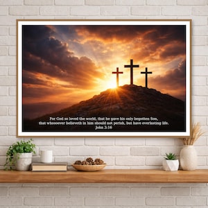 Puede incluir: Impresión enmarcada con tres cruces silueteadas contra un atardecer dramático. El cielo está lleno de tonos naranjas y amarillos, con texto de Juan 3:16 en la parte inferior. La obra de arte se exhibe en una estantería de madera contra una pared de ladrillos blancos.