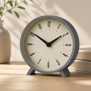 May include: A gray, round analog clock with a cream-colored face. Black numerals and hands mark the time. The clock has a simple, modern design and sits on a wooden surface. The clock is approximately 5 inches in diameter.