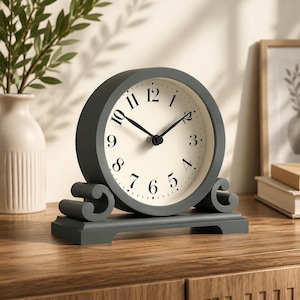 May include: A gray, round mantel clock with a cream-colored face and black hands and numerals. The clock has a decorative base with scroll details. The clock is sitting on a wooden surface.