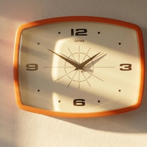 May include: A retro-style wall clock with an orange frame. The clock face is off-white with black numerals at 3, 6, 9, and 12 o'clock. The clock hands are black, and the word "Jones" is printed above the 12. The clock has a starburst design.