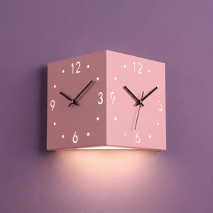 May include: A pink, cube-shaped wall clock with three visible faces, each displaying the numbers 12, 3, 6, and 9 in white. The clock has black hands and white dot hour markers. A warm light emanates from the bottom.