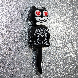 May include: A black Kit-Cat Klock with a cat face, white bow tie, and red heart eyes. The clock has a white clock face with black numbers and hands. The clock's tail is long and curved.