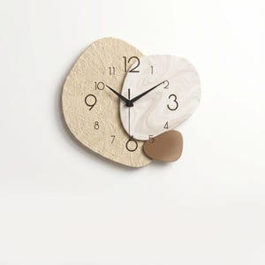 May include: A modern wall clock with a unique design. The clock features a textured beige background, a white and beige marbled section, and a small brown accent. Black hands and numerals complete the design. The clock measures approximately 12 inches in diameter.