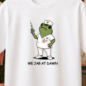 Può includere: Una t-shirt bianca a maniche corte con la grafica di una rana verde vestita da infermiere, che indossa un cappellino bianco con una croce rossa e tiene in mano una siringa. Sotto la rana, il testo recita WE JAB AT DAWN in lettere nere.