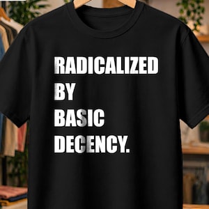 Può includere: Una t-shirt nera a maniche corte con la scritta bianca RADICALIZED BY BASIC DECENCY stampata in un carattere sans-serif in grassetto sul davanti. Il capo è appeso a una gruccia di legno su uno sfondo sfocato di scaffali di abbigliamento.