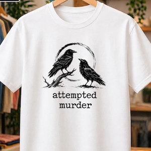 Pode incluir: Uma t-shirt branca de manga curta com um gráfico preto de dois corvos pousados num ramo dentro de um contorno circular. Abaixo da ilustração, o texto diz attempted murder numa fonte preta com estilo de máquina de escrever.