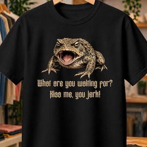 Può includere: Una t-shirt nera a maniche corte con la grafica di un rospo marrone a bocca aperta. Sotto il rospo, il testo recita: What are you waiting for? Kiss me, you jerk! La maglietta è esposta su una gruccia di legno davanti a un appendiabiti.