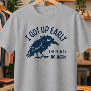 Könnte beinhalten: Ein graues, meliertes Kurzarm-T-Shirt mit einer dunkelblauen Grafik eines Raben und dem Text "I GOT UP EARLY THERE WAS NO WORM" in einer abgenutzten Schriftart. Das T-Shirt hat einen Rundhalsausschnitt und hängt an einem Holzkleiderbügel.