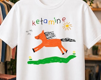May include: A white short-sleeve t-shirt featuring a colorful graphic of an orange horse running on a green patch of grass. Above the horse, the word ketamine is printed in multicolored letters, accompanied by a yellow sun and small birds.