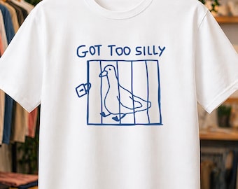 May include: A white short-sleeved t-shirt featuring a blue graphic of a duck inside a cage with a padlock. Above the cage, the text reads "GOT TOO SILLY" in blue capital letters. The shirt is displayed on a wooden hanger against a retail background.