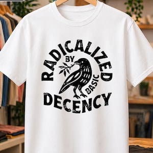 Può includere: Una t-shirt bianca a maniche corte con una stampa grafica nera di un uccello che tiene un piccolo ramo. Il testo RADICALIZED BY BASIC DECENCY è disposto in un motivo circolare attorno all'illustrazione in un carattere nero dall'aspetto vissuto.