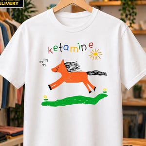 May include: A white short-sleeved t-shirt featuring a colorful graphic print of an orange horse running above a green patch of grass. The text "ketamine" is printed in multicolored letters above the horse, accompanied by a yellow sun illustration.