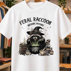 Può includere: Una t-shirt bianca a maniche corte con la grafica di un procione che indossa un cappello da strega e mescola un calderone ribollente. Il testo sopra recita FERAL RACCOON BREWING POTIONS. Il design include libri, un teschio e varie boccette.