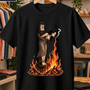 Puede incluir: Una camiseta negra con una ilustración gráfica de una persona con cabello largo y oscuro atada a un poste de madera, vistiendo un vestido sencillo, mientras llamas surgen de la base y humo sale de un cigarrillo en su boca.