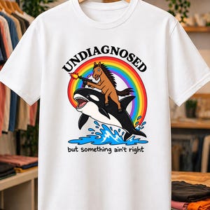 Könnte beinhalten: Ein weißes Kurzarm-T-Shirt mit einer Grafik eines braunen Pferdes, das auf einem schwarz-weißen Orca reitet. Das Pferd hält eine Schusswaffe mit Mündungsfeuer. Ein Regenbogen wölbt sich über blauem Spritzwasser. Der Text lautet UNDIAGNOSED but something ain't right.