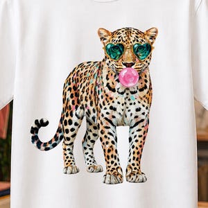 Può includere: Una t-shirt bianca a maniche corte con una stampa grafica di un leopardo in piedi di profilo. Il leopardo ha il manto maculato, indossa occhiali da sole a forma di cuore color verde acqua e soffia una grande bolla rosa dalla bocca.