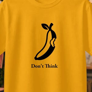Può includere: T-shirt giallo senape con grafica nera di una banana parzialmente mangiata e la scritta "Don't Think". La maglietta è appesa a una gruccia di legno. Altre magliette sono visibili sullo sfondo.