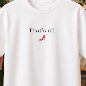 Puede incluir: Camiseta blanca con la frase "That's all." y un dibujo de un zapato de tacón rojo. La camiseta está colgada en una percha de madera. Un banner publicitario con una furgoneta de reparto es visible en la esquina superior izquierda.