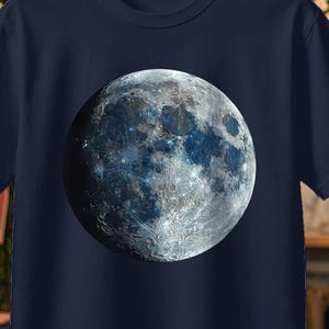 Può includere: T-shirt blu navy con un'immagine dettagliata della luna. La grafica della luna è un mix di blu, grigio e bianco, con crateri e texture visibili. La maglietta è appesa a un appendiabiti di legno. Il testo "BUY X3 FREE DELIVERY" è in alto a sinistra.