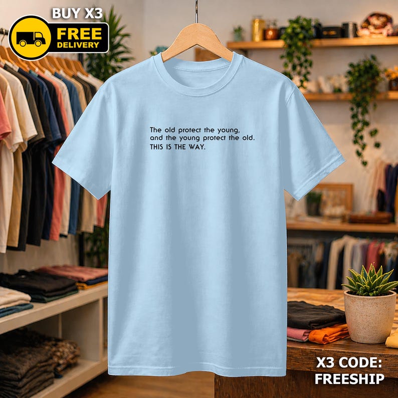 May include: A pale blue t-shirt featuring the text "The old protect the young, and the young protect the old. THIS IS THE WAY." The shirt is hanging on a wooden hanger. The image also includes a "BUY X3 FREE DELIVERY" graphic.