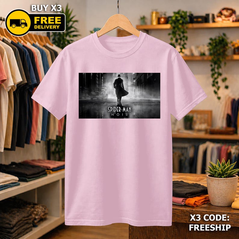 May include: A light pink short-sleeved t-shirt featuring a rectangular graphic print of a silhouetted figure in a trench coat and fedora walking down a dark, foggy street with the text Spider-Man Noir printed in white below the figure.