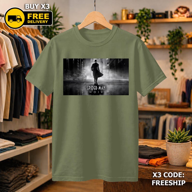 May include: An olive green short-sleeve t-shirt featuring a rectangular black and white graphic print on the chest. The graphic depicts a silhouette of a figure in a trench coat and hat standing in a foggy street with the text Spider-Man Noir.