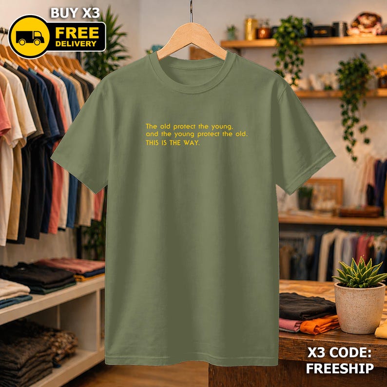 May include: An olive green t-shirt with the text "The old protect the young, and the young protect the old. THIS IS THE WAY." in yellow. The shirt is hanging on a wooden hanger.
