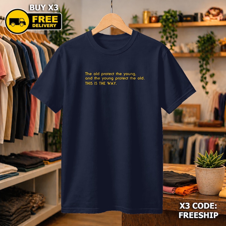 May include: A navy blue t-shirt with yellow text: "The old protect the young, and the young protect the old. THIS IS THE WAY." The shirt is displayed on a wooden hanger. The image also includes a "BUY X3 FREE DELIVERY" graphic.