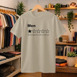 May include: A beige short-sleeved t-shirt featuring the black text 'Men' above a five-star rating graphic with one filled-in star and four empty stars, followed by the text 'Absolute nightmares, would not recommend.' printed in black.