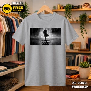 May include: A heather gray short-sleeve t-shirt featuring a rectangular black and white graphic print of a silhouetted figure in a trench coat and fedora walking down a city street with the text Spider-Man Noir printed below the figure.