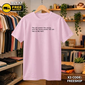 May include: A pale pink t-shirt with the text "The old protect the young, and the young protect the old. THIS IS THE WAY." The shirt is displayed on a wooden hanger. The image also includes a "BUY X3 FREE DELIVERY" graphic.