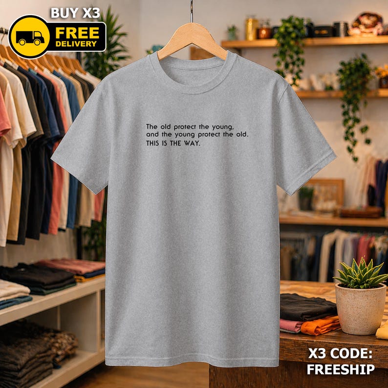 May include: A grey t-shirt with the quote "The old protect the young, and the young protect the old. THIS IS THE WAY." The shirt is displayed on a wooden hanger. The t-shirt is part of a clothing collection.