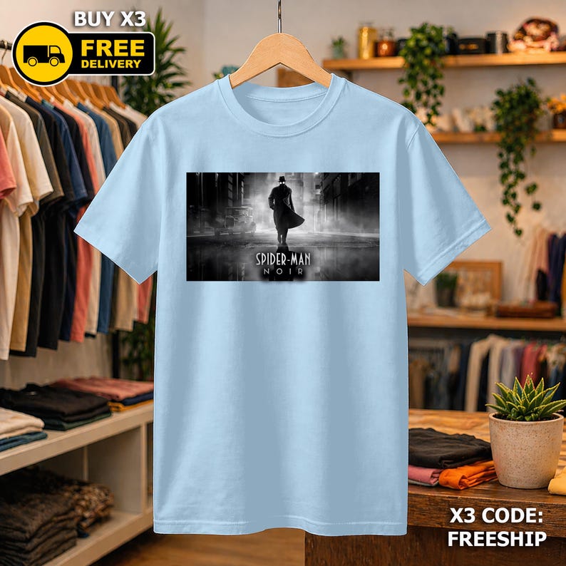 May include: A light blue t-shirt featuring a rectangular graphic print of a silhouetted figure in a trench coat walking down a foggy street. The text Spider-Man Noir is printed in white below the figure. The shirt is displayed on a wooden hanger.